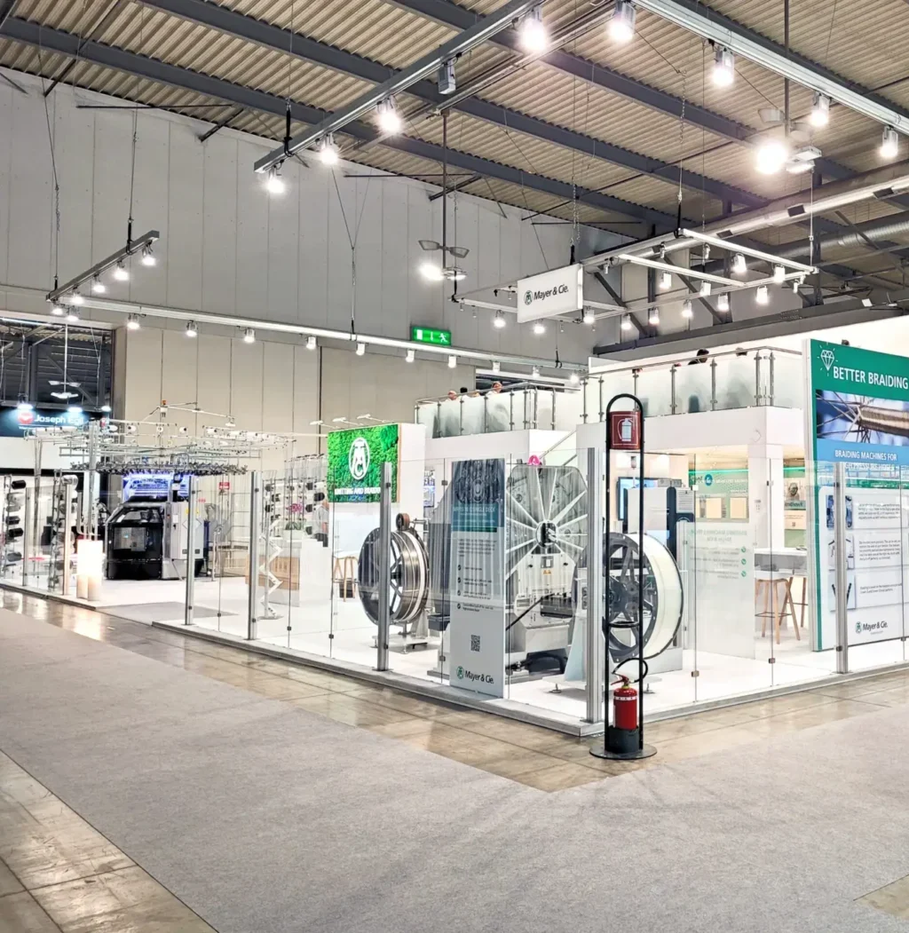Großer Doppelstock-Messestand mit einer Fläche von 400 qm und einer zweiten Ebene von 100 qm. Offenes, modernes Design mit Glaswänden, großzügigen Ausstellungsflächen und markantem Branding der Firma Mayer & Cie. Der Stand zeigt industrielle Maschinen, informative Displays und Sitzbereiche auf beiden Ebenen. Helle Beleuchtung und klare Strukturen laden Besucher ein.