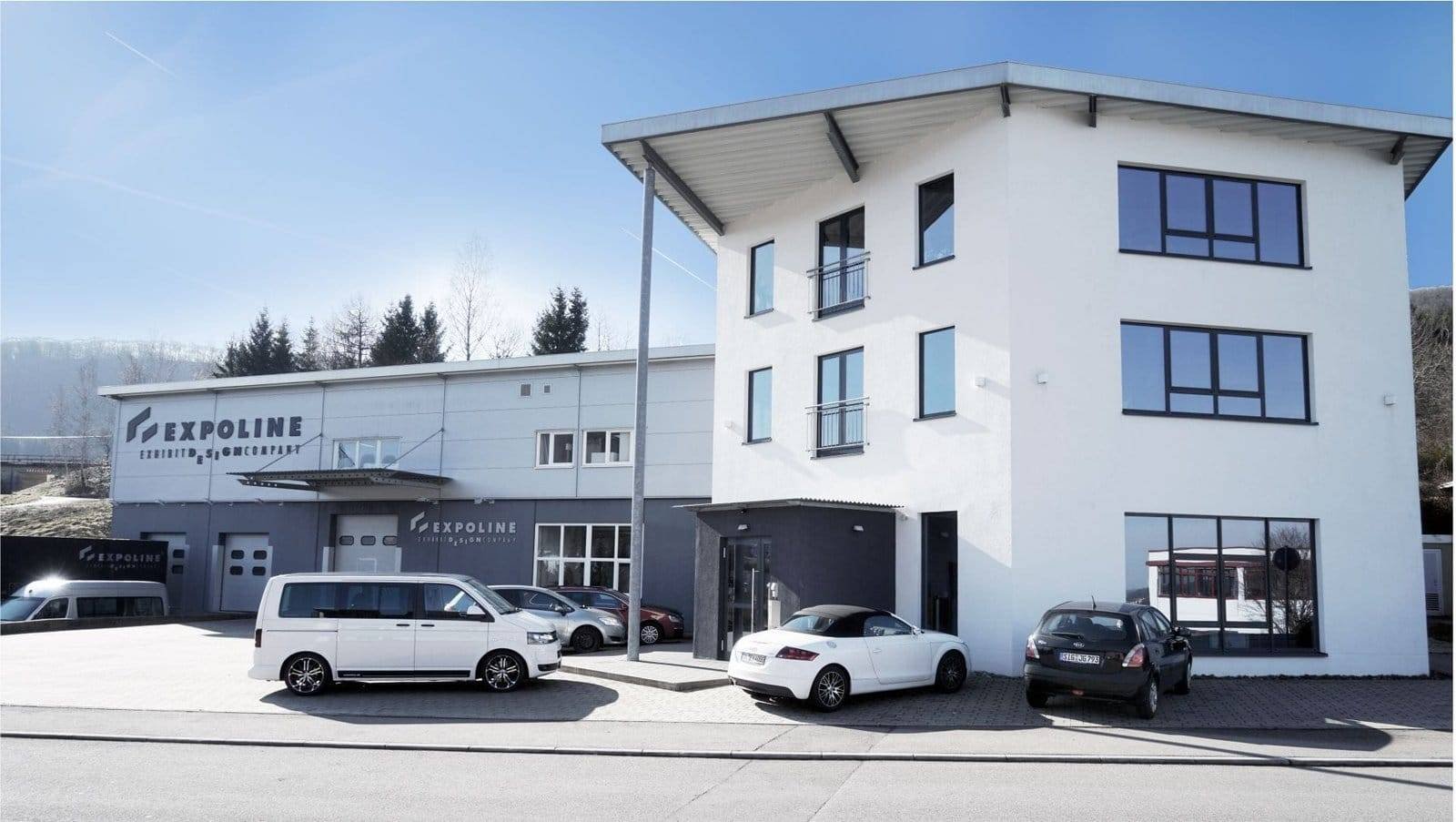 Firmengebaeude albstadt | expoline messedesign Außenansicht des expoline-firmensitzes mit modernem bürogebäude, produktionshalle und parkplatz im vordergrund bei klarem himmel.