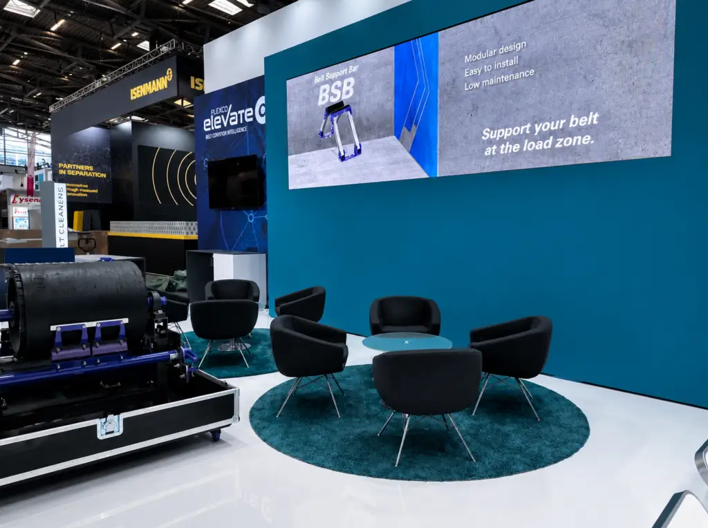 Exhibition booth with a teal wall, six black chairs around a round teal rug and glass coffee table; large display screens overhead and industrial equipment to the left.