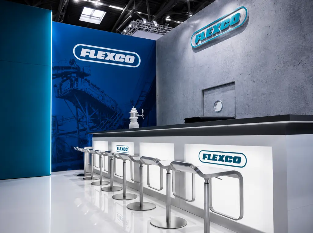 Flexco_Startbild • EXPOLINE Messedesign • EXPOLINE Messedesign FLEXCO trade-show booth with a blue mural, illuminated logo, and white bar counter with chrome stools under bright lighting.