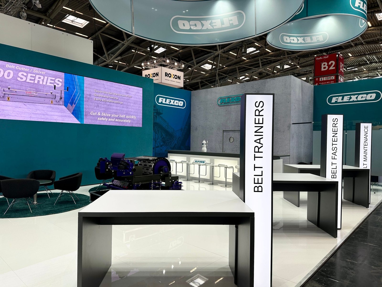 Moderner Messestand der Firma FLEXCO mit weiß-türkisem Design, großem Logo, Multimedia-Bildschirm und ausgestellten technischen Produkten. Präsentationsflächen mit klaren Linien zeigen Förderbandlösungen und Komponenten. Hängende Deckenbanner und prominente Kontaktdaten runden das professionelle Erscheinungsbild ab.
